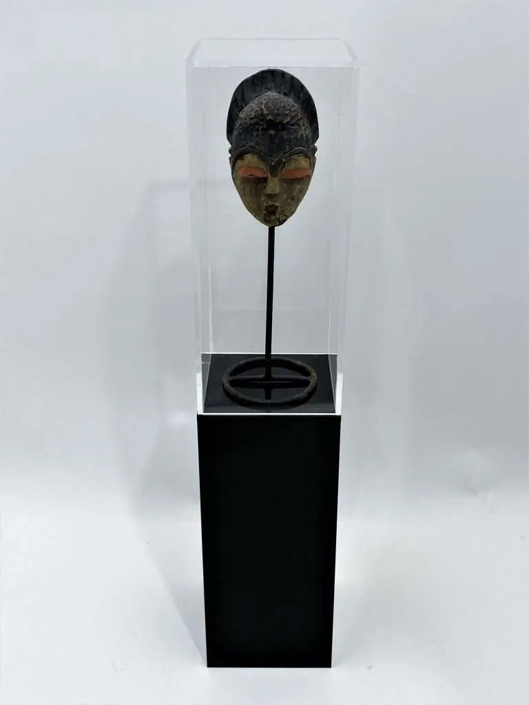 Clear acrylic display case atop a black acrylic pedestal presenting a carved wood mask on a stand.